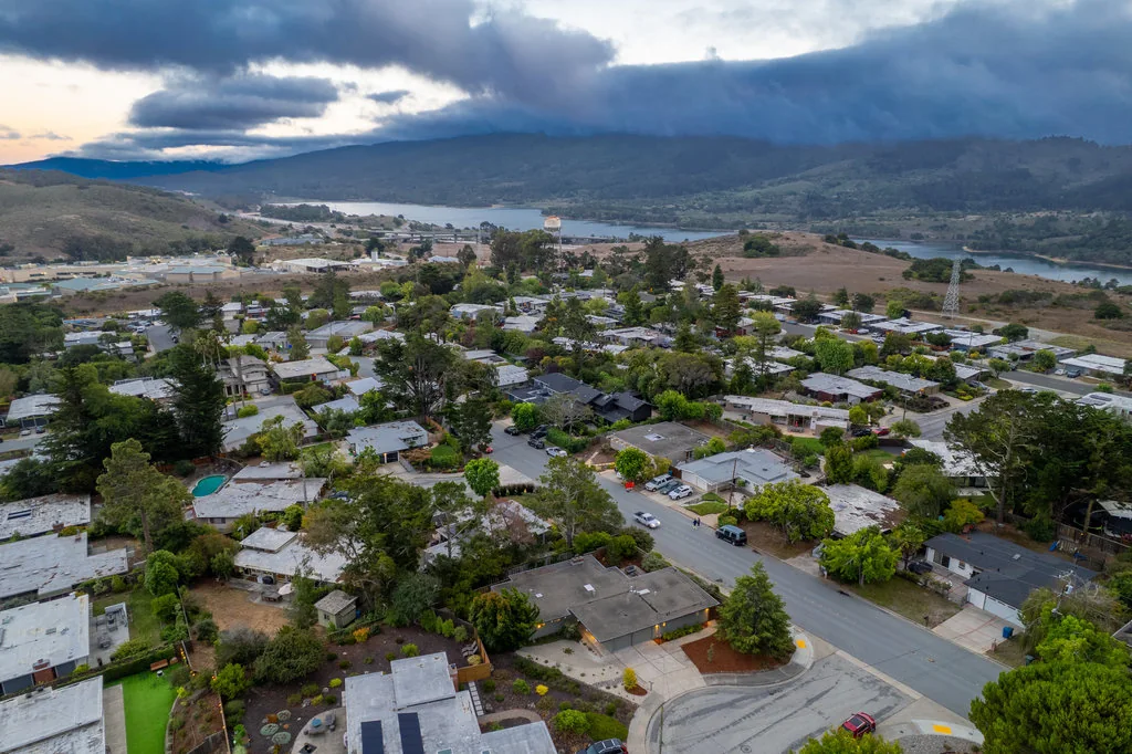 An aerial photograph at dusk over the San Mateo Highlands: flat-roofed Eichlers on curving streets, with Crystal Springs Reservoir and the undeveloped watershed ridge beyond.
