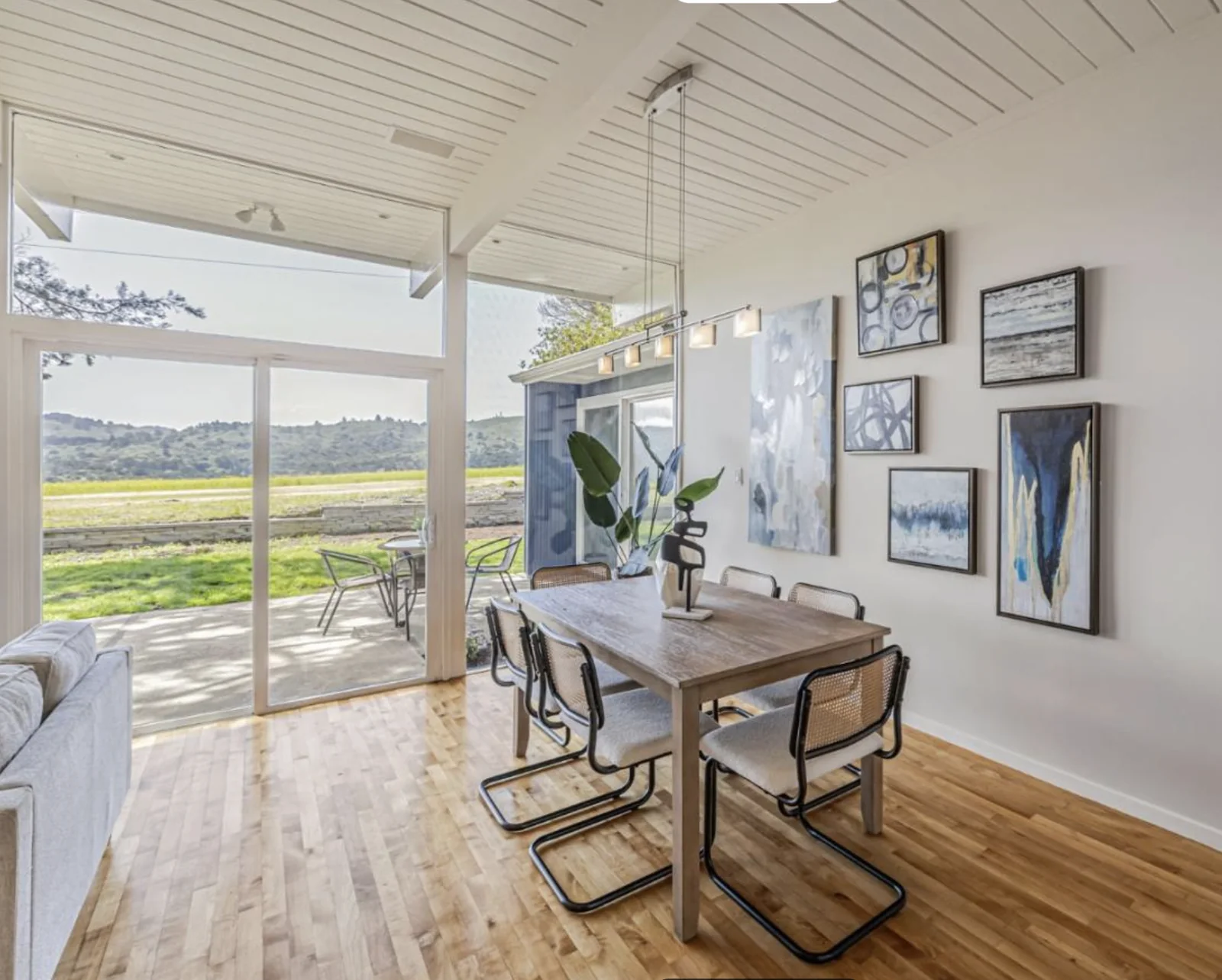 A dining area with a gallery wall of abstract art, a linear pendant over the table, and a sliding glass door to the patio and meadow.