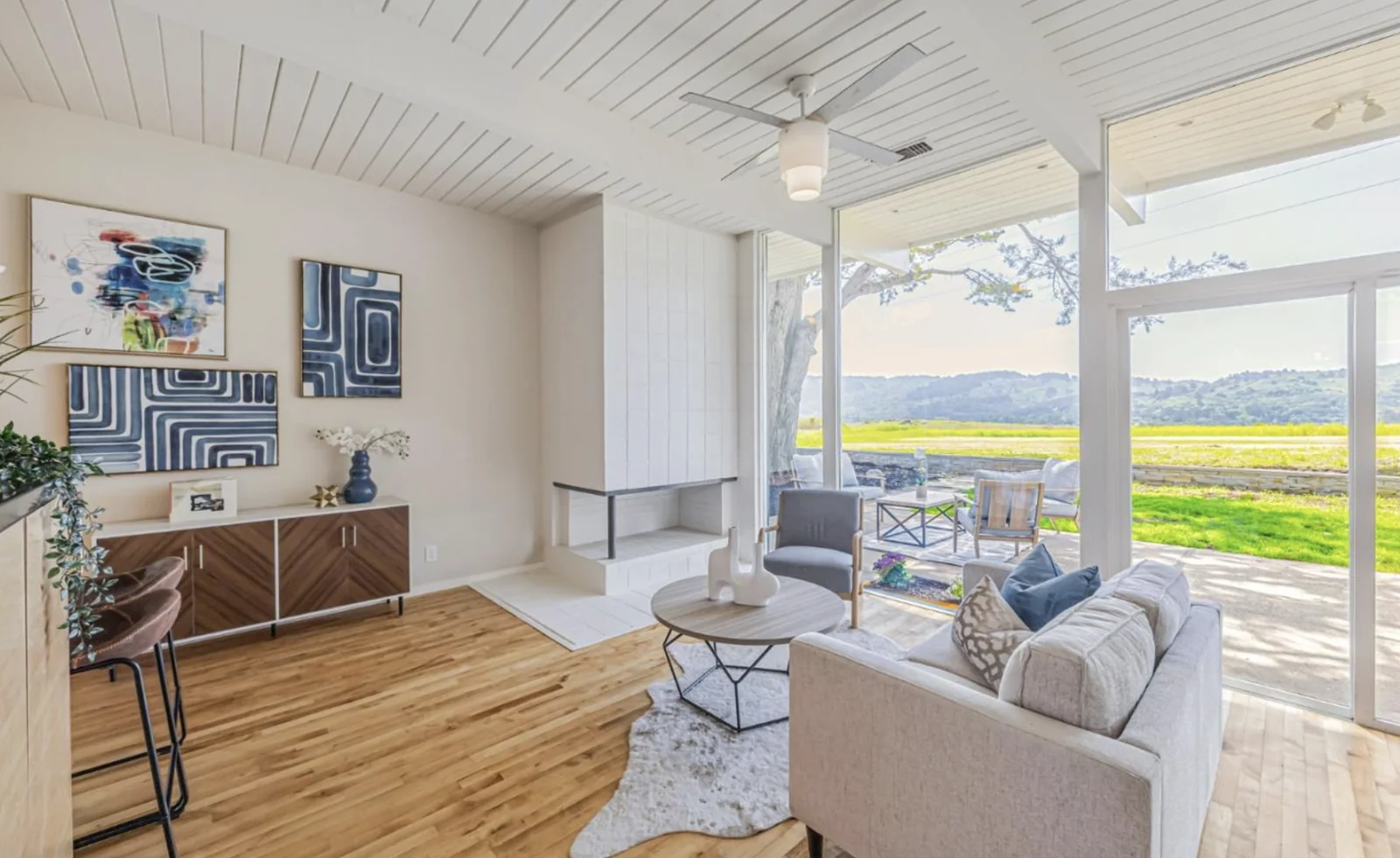 A living room with a white tiled fireplace, exposed beams in a white tongue-and-groove ceiling, and full-height glass looking onto open meadow and wooded hills.