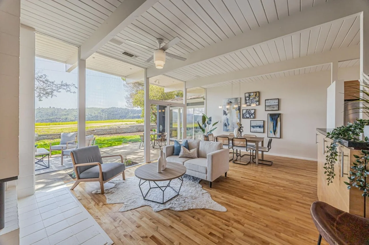 The living room of a San Mateo Highlands Eichler: white tongue-and-groove ceiling on exposed beams, oak floors, and a full-height glass wall opening onto a terrace and open grassland beyond.