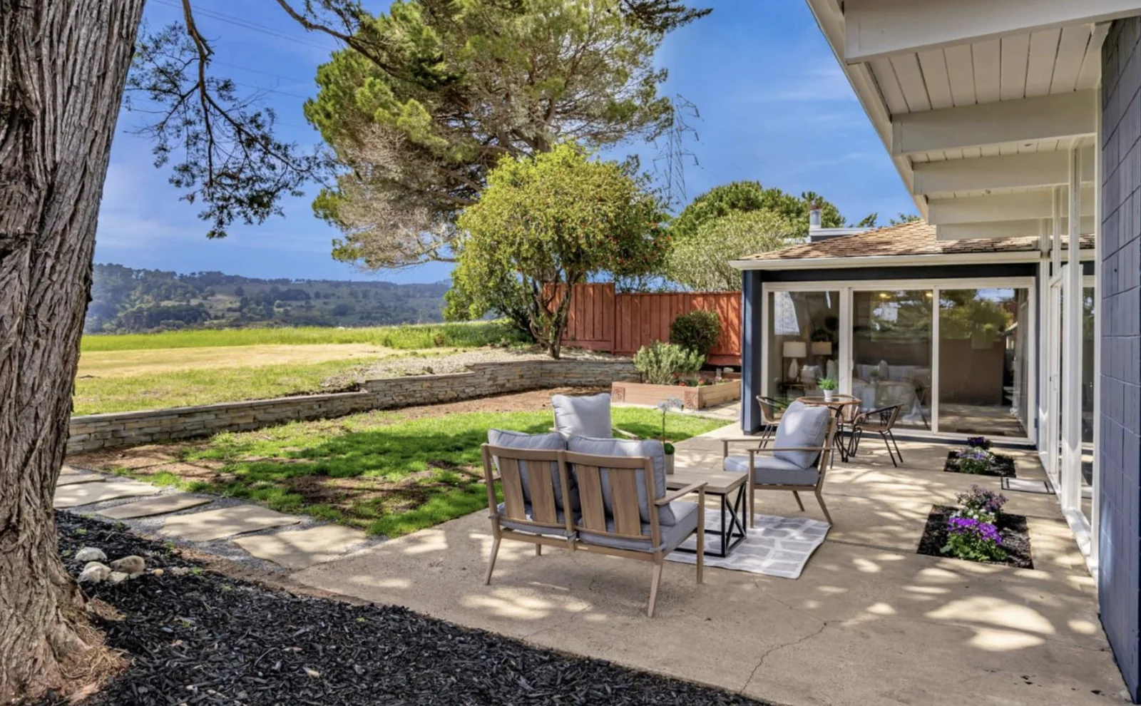The patio from under the cypress, looking along the glass wall of the house to the right and out over the meadow and hills to the left.