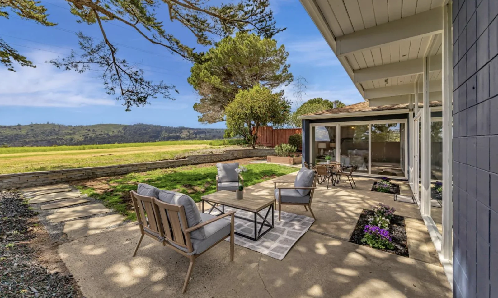 A concrete patio with cushioned seating beside a wall of sliding glass, looking across a low stone retaining wall to a wide meadow and wooded hills.