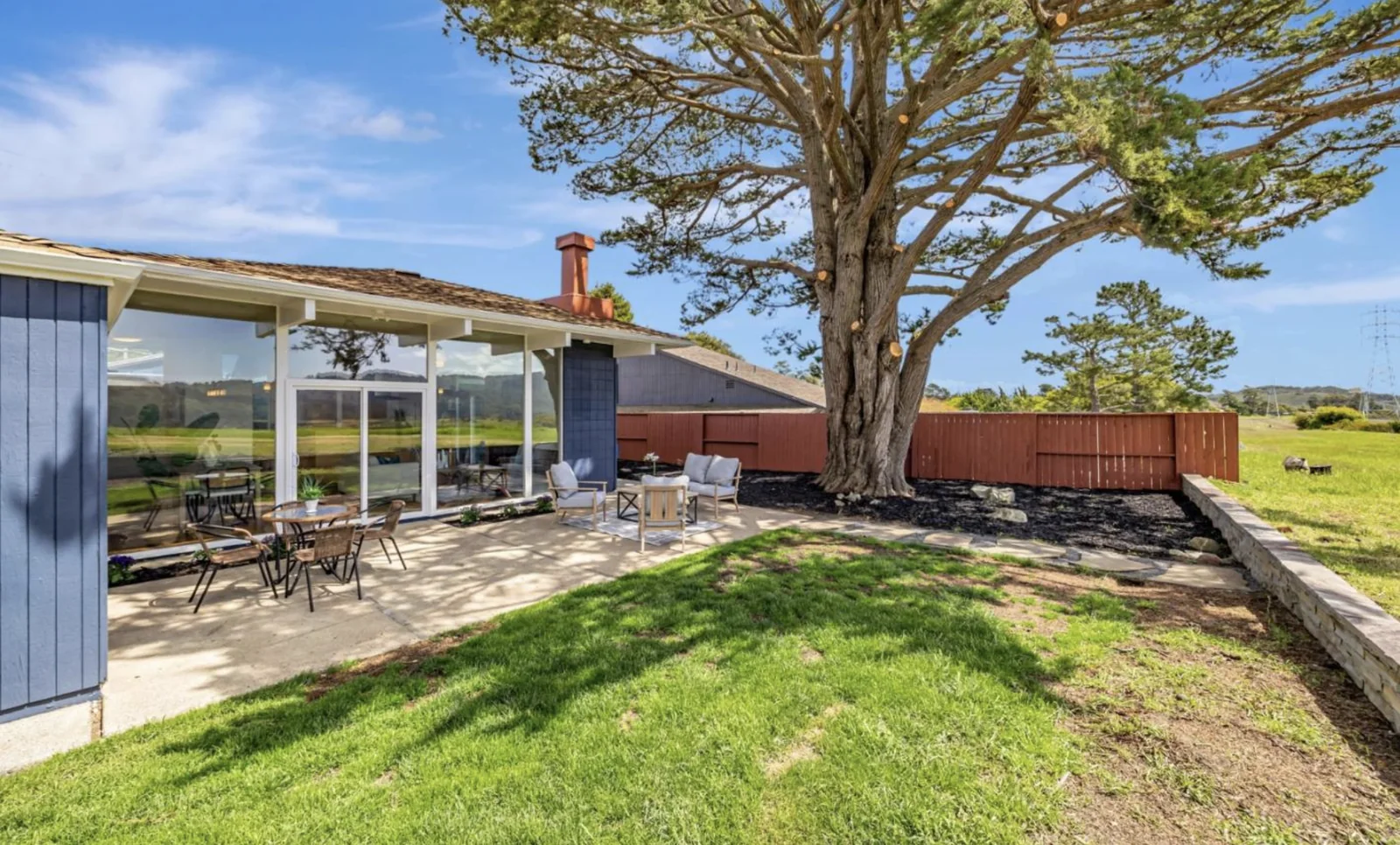 The side and rear of the house seen past a very large Monterey cypress, with a red-stained fence marking the property line and open meadow immediately beyond it.