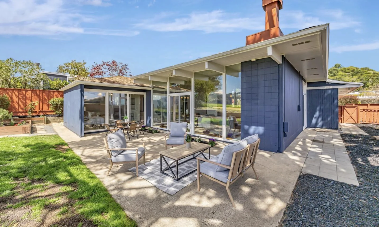 The rear elevation of an Eichler with a low shed roof in wood shakes, deep eaves, floor-to-ceiling glass walls, and a navy-painted concrete-block chimney mass carrying a terracotta flue.