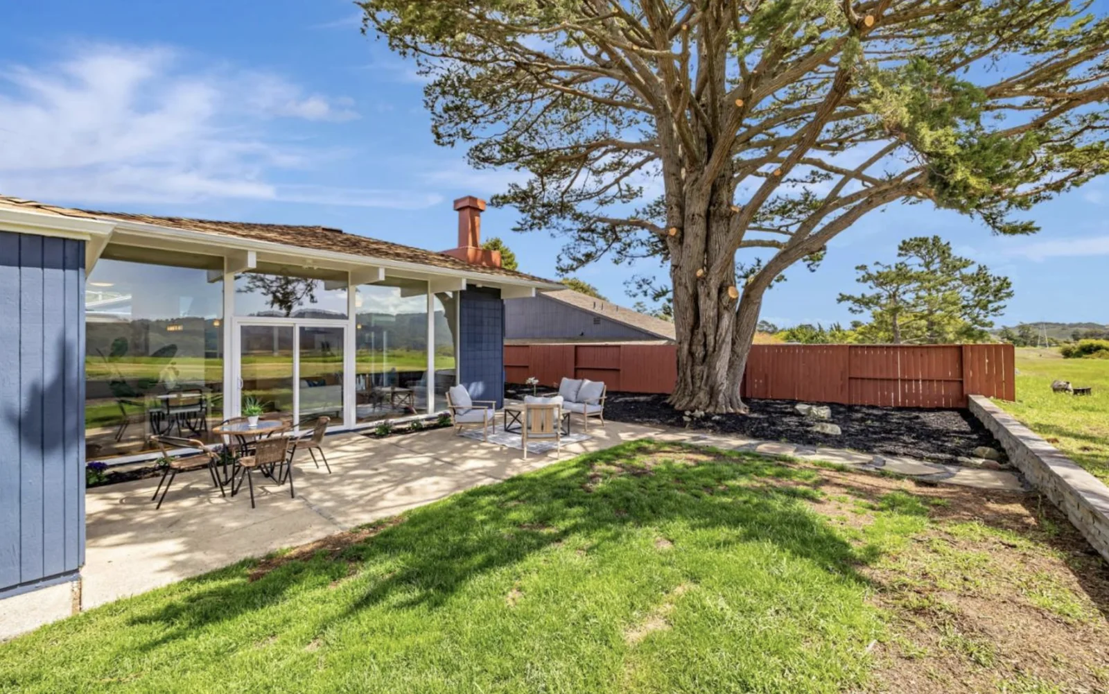 A view along the glass wall toward the navy chimney mass, with the cypress overhead and the meadow visible both through the house and beside it.