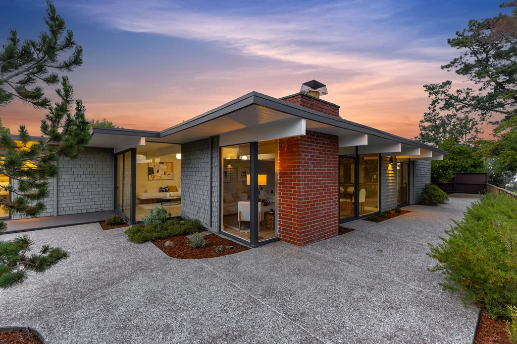 The private side of an Eichler at dusk: a red brick chimney mass flanked by full-height glass walls, with the interior lit and an aggregate terrace in front.