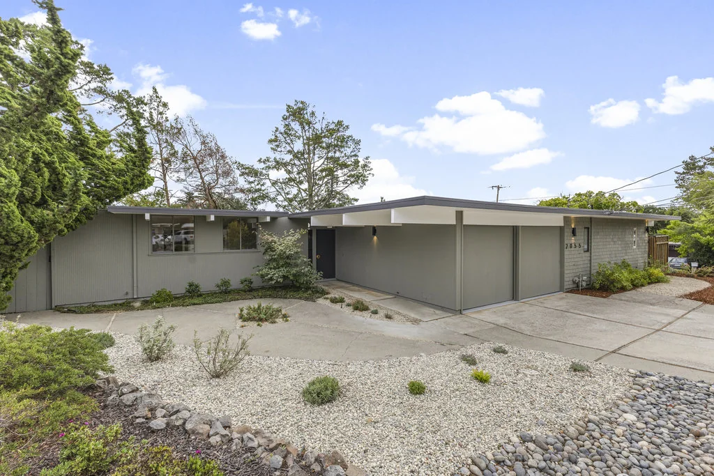 A daylight three-quarter view of an Eichler entry: a low folded roof with beam ends projecting past the wall, a recessed entry slot, and the ground running straight to the door with no step.