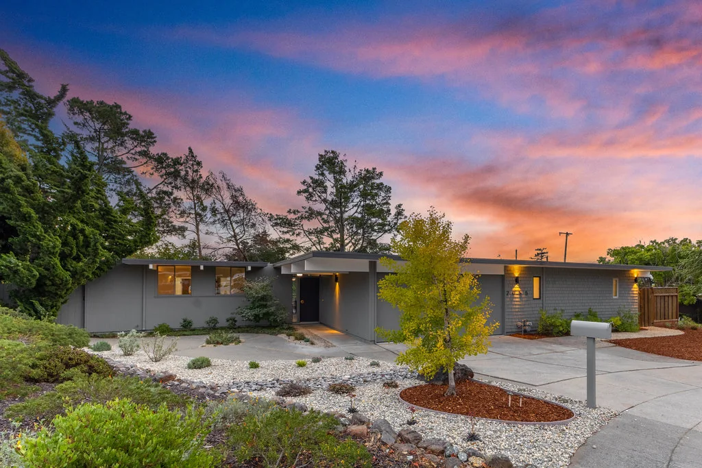 A single-storey Eichler on Ticonderoga Drive in the San Mateo Highlands at dusk, its low roof folding over a recessed entry, with a drought-tolerant gravel and boulder front garden.