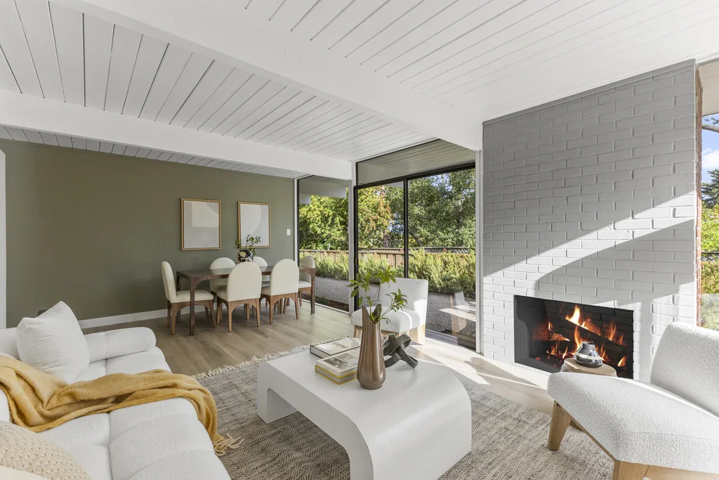 An Eichler living room with a painted brick fireplace, exposed beams in a white tongue-and-groove ceiling, and an olive dining wall with glass to the garden beyond.
