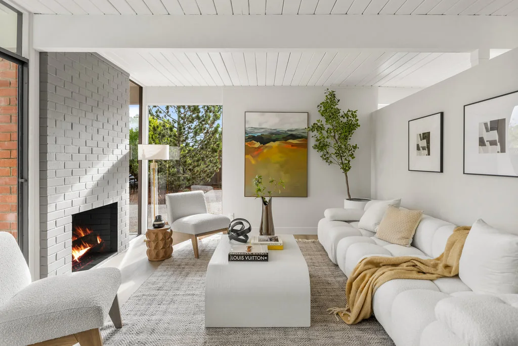 A living room with a white-painted brick fireplace, exposed beams in a white tongue-and-groove ceiling, and floor-to-ceiling glass.