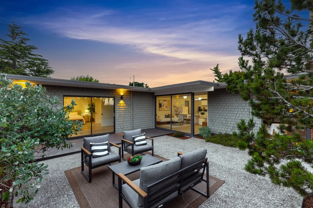 A rear patio at dusk with modern outdoor seating, the house glowing from inside through two sliding glass walls, one to a bedroom and one to the living room.