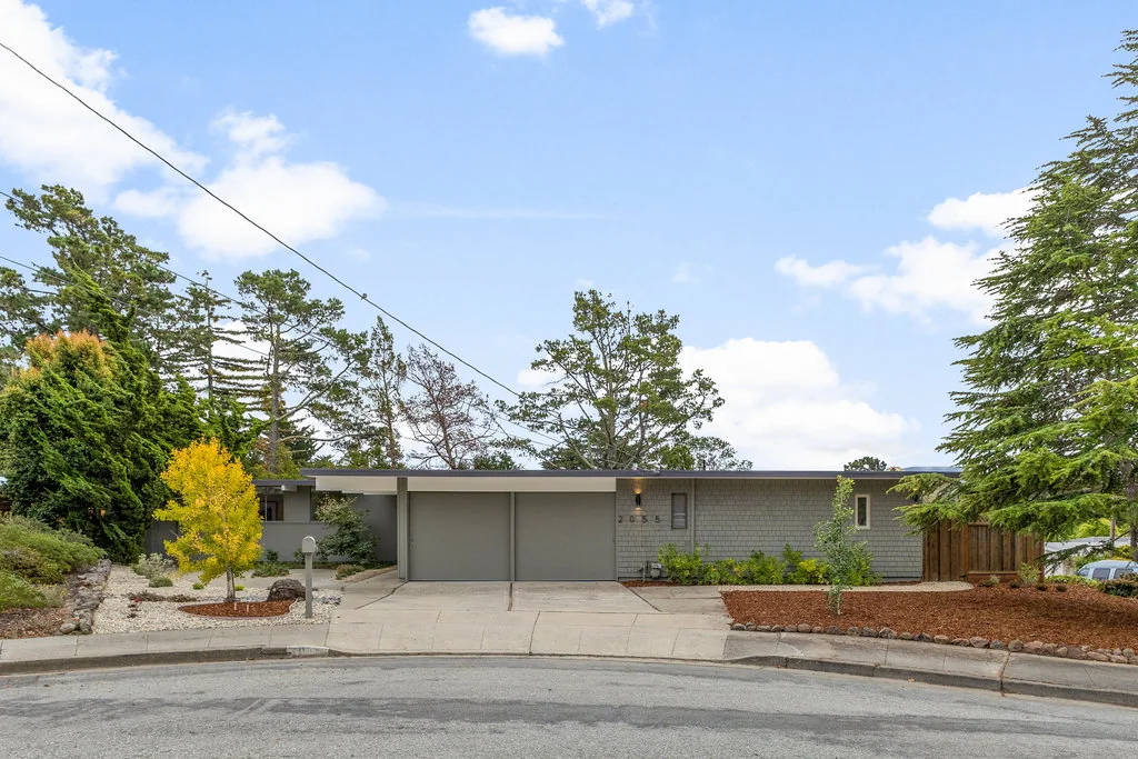 A Highlands Eichler seen straight on from across a cul-de-sac: a long low roofline, paired garage doors, textured block and vertical board cladding, mature conifers behind.