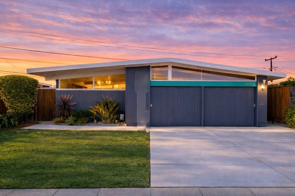 A 19th Avenue Park Eichler at sunset: a low gabled roof with deep overhangs, dark blue vertical board siding, a teal panel above twin garage doors carrying the number 637, and clerestory glass under the ridge.
