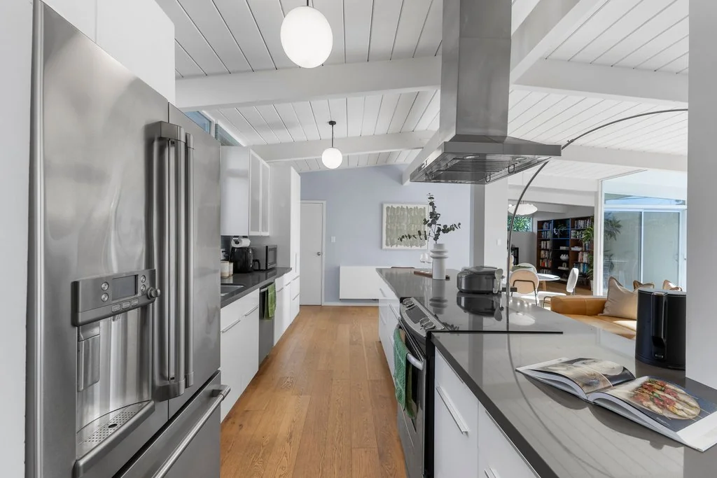 A galley kitchen with white cabinets, dark counters, a stainless island hood, globe pendants and a white-painted beamed ceiling running the length of the house.
