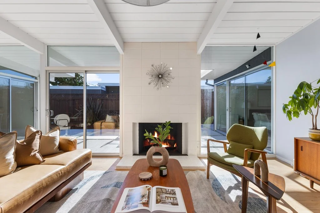 A living room with a white concrete-block fireplace at its centre, exposed beams in a white tongue-and-groove ceiling, and sliding glass walls opening to outdoor space on both sides.