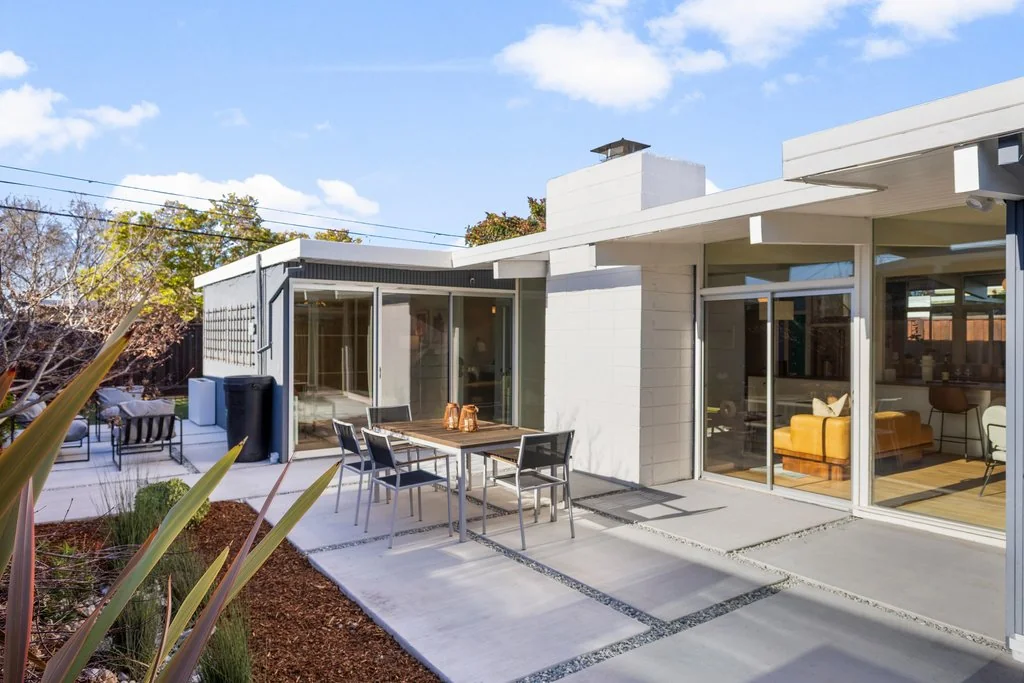 A rear patio of concrete pavers with a dining table, enclosed by the L of the house with a white concrete-block chimney mass and walls of sliding glass.