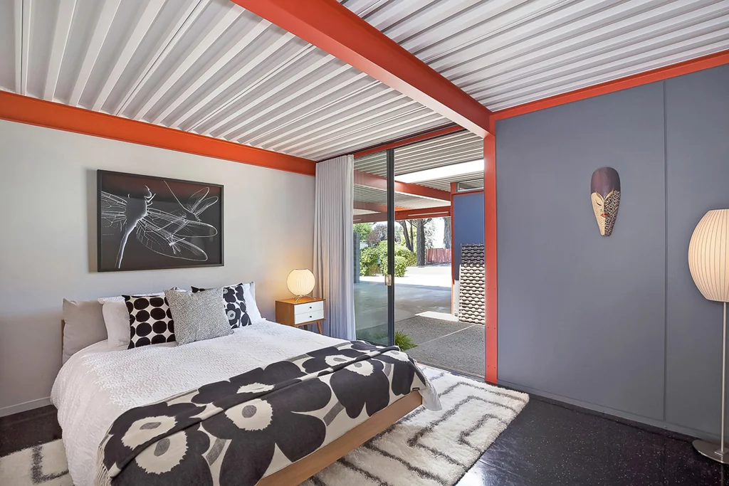 A bedroom with white and grey walls, an orange steel beam overhead, and a sliding glass door opening onto a covered walkway.