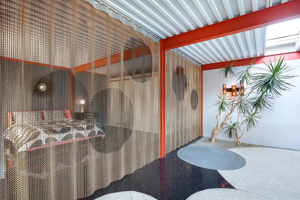 A bedroom enclosed by floor-to-ceiling metal chain-mail curtains with circular motifs, behind an orange steel column, with a dracaena in a planted bed beside it.