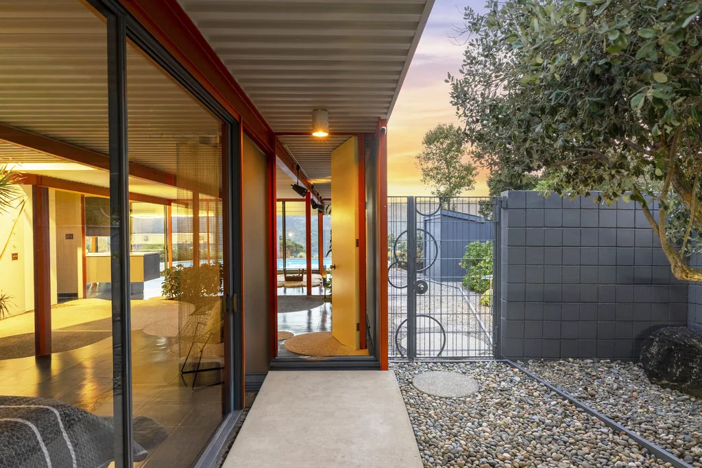 The entry walk at dusk: a concrete path beside a glazed wall, a yellow front door standing open, a circular-motif metal gate and a screen-block wall with a river-stone bed.