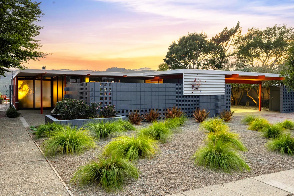 The X-100 at 1586 Lexington Avenue at sunset: a long flat roof carried on orange-painted steel, a grey screen-block wall with the house number 1586, and a gravel garden of grasses.