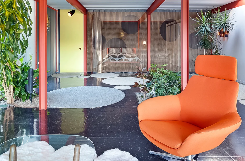 An orange lounge chair on a polished black terrazzo floor inlaid with pale circles, looking through planting to the chain-mail-curtained bedroom beyond.