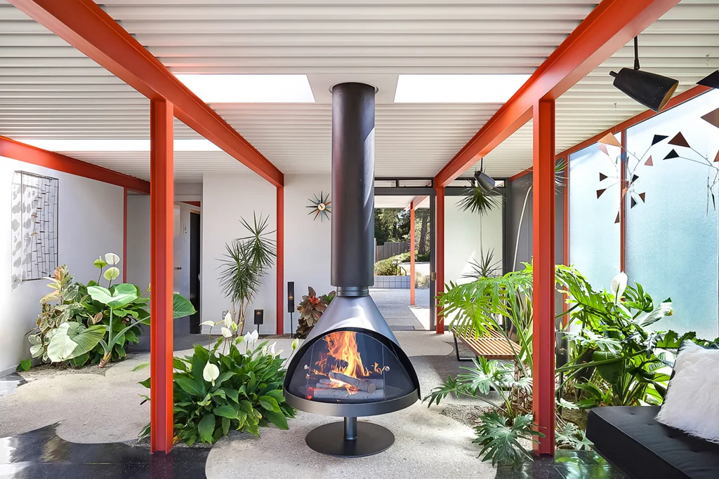 An interior planted garden under a skylight, framed by orange-painted steel columns and a white corrugated steel roof deck, with a free-standing black cone fireplace at the centre and tropical planting all around.