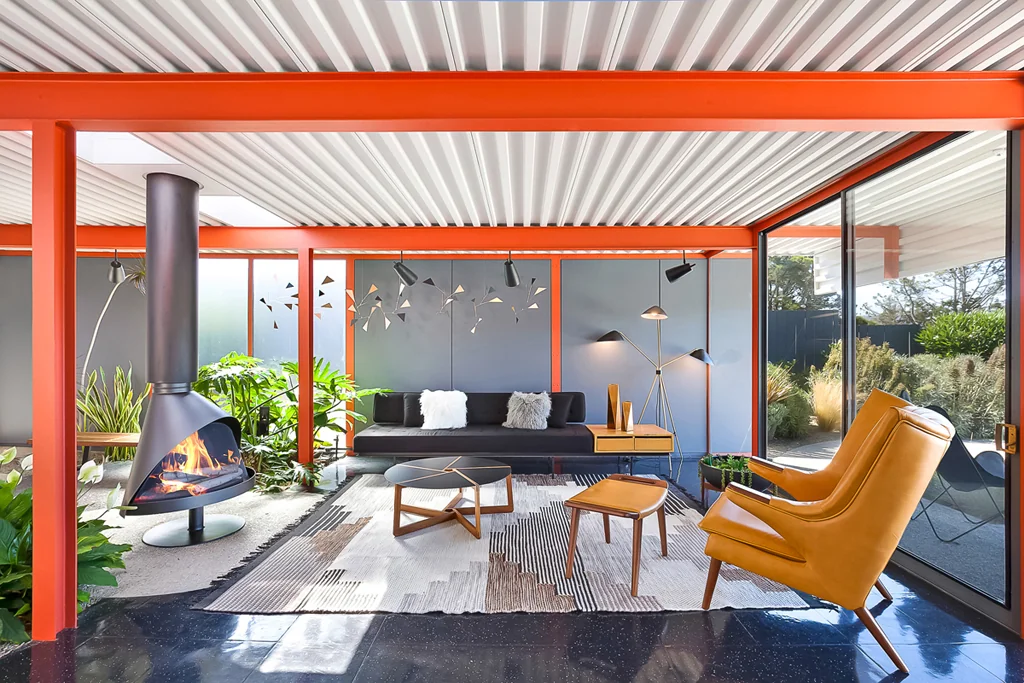 A seating area inside the planted garden with a cone fireplace alight, orange steel columns, a low black sofa and a metal mobile on the wall behind.