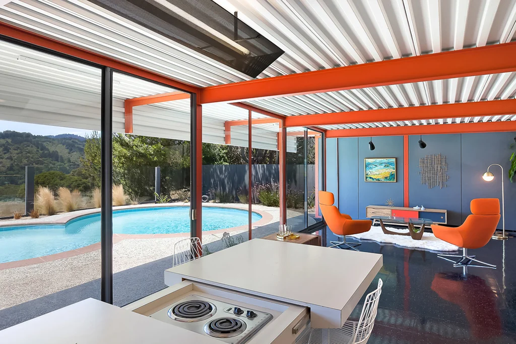 A close view of a white kitchen table with an electric cooktop recessed into its surface, with the pool and living room visible beyond through full-height glass.