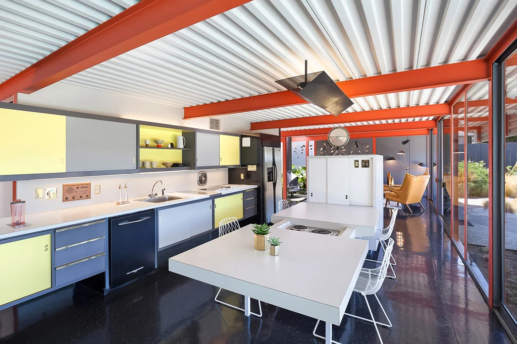 A kitchen with pale yellow and grey cabinet fronts, a long counter, and two white freestanding islands under an orange steel beam and white corrugated ceiling.