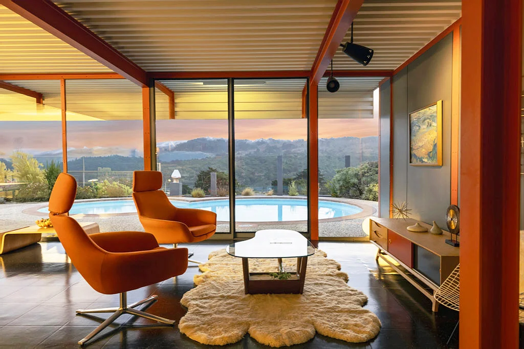 A living room with two orange swivel chairs, orange steel columns and beams, a white corrugated steel ceiling, and full-height glass looking onto a kidney-shaped pool with hills beyond at dusk.