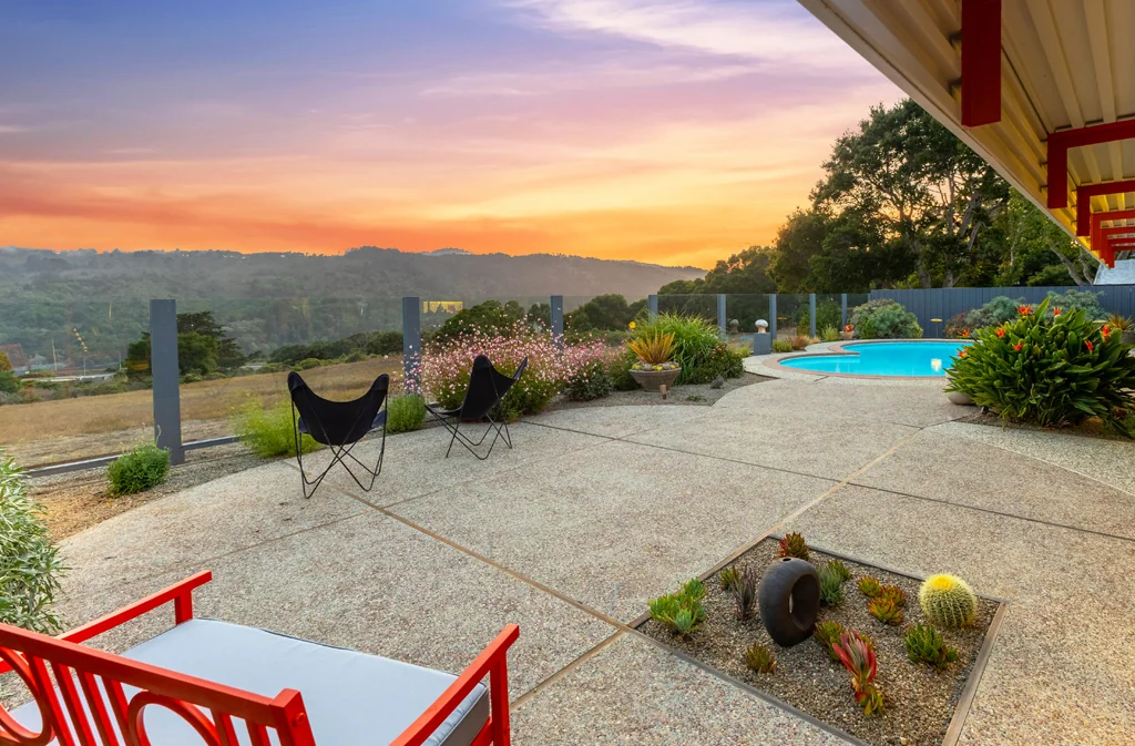 A wide aggregate terrace with butterfly chairs and a pool at sunset, looking west over open grassland to wooded ridges.