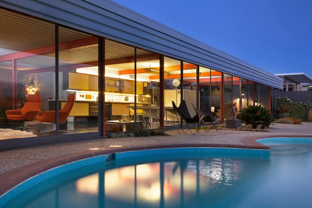 The house at dusk from across the pool, the whole glazed wall lit from within showing the kitchen and living room, with the water reflecting the light.