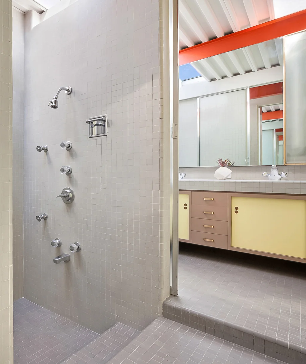 A tiled shower with one overhead head and five body sprays on the wall, beside a vanity with pale yellow cabinet fronts lit by a skylight.