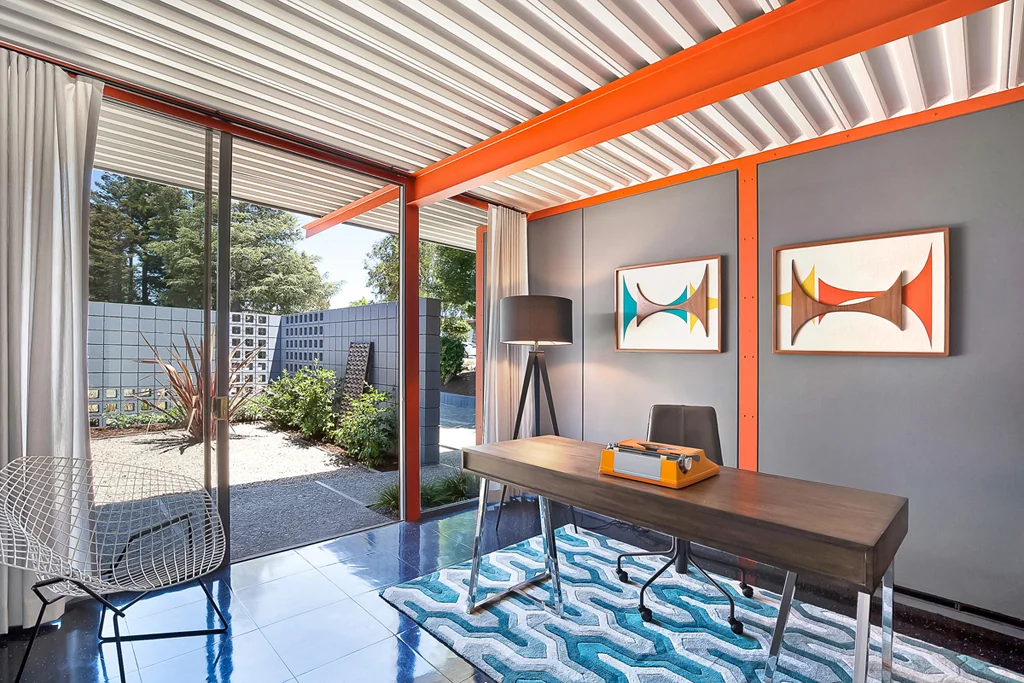 A study with a desk, two abstract mid-century artworks on a grey wall, an orange steel beam, and sliding glass onto a screen-block courtyard.